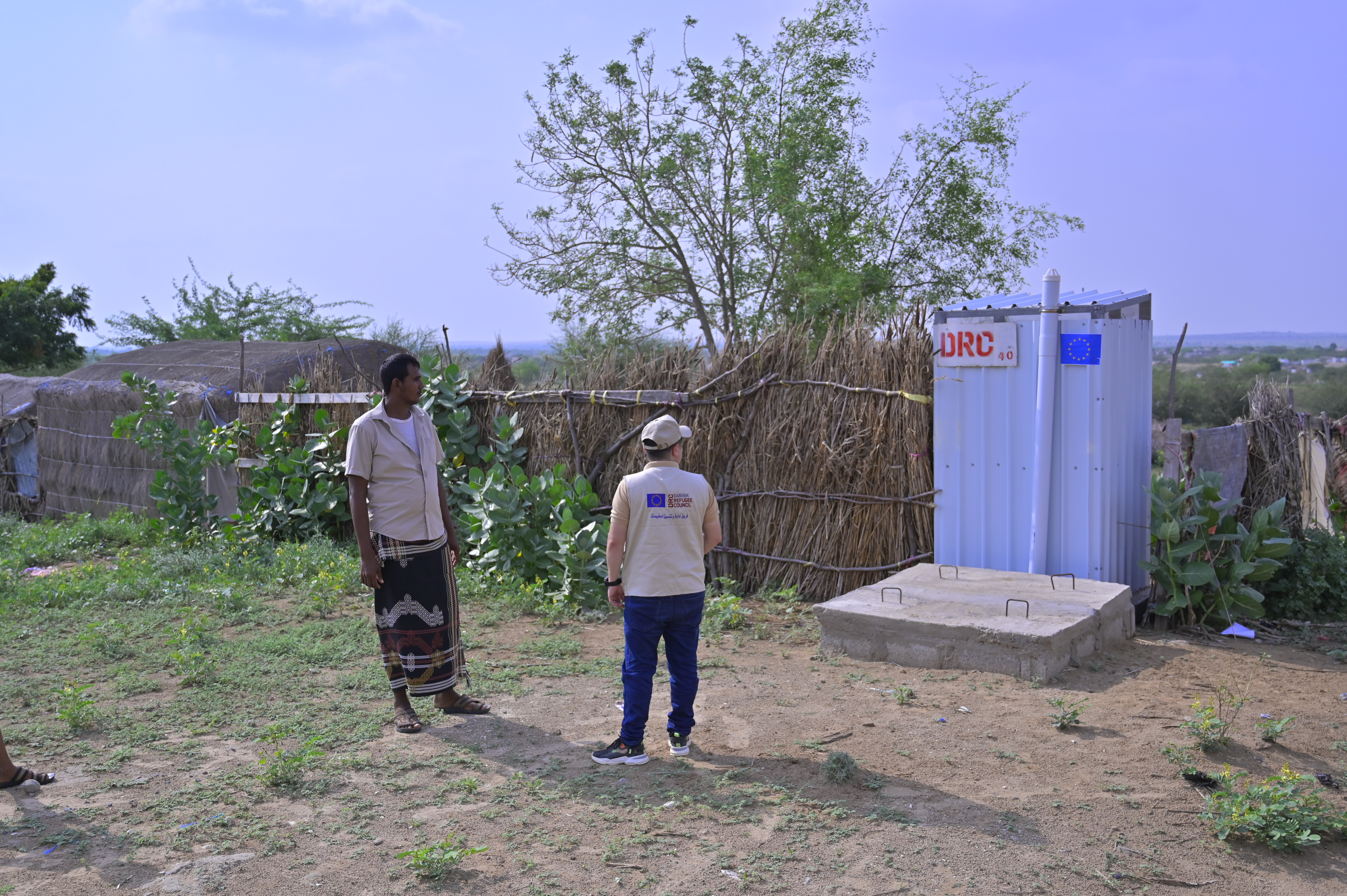Mohammed, a DRC Water, Sanitation and Hygiene (WASH) Assistant, with Hussein in front of a latrine installed by DRC at Al-Radha Camp, Hajjah Governorate, September 2025. Photo by: Reyad Al Qudami